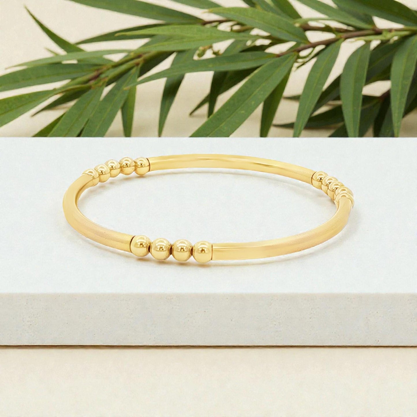 A gold-colored bracelet with a row of small, round beads on it, placed on a white surface with green leaves in the background.