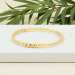 A gold-colored bracelet with a row of small, round beads on it, placed on a white surface with green leaves in the background.