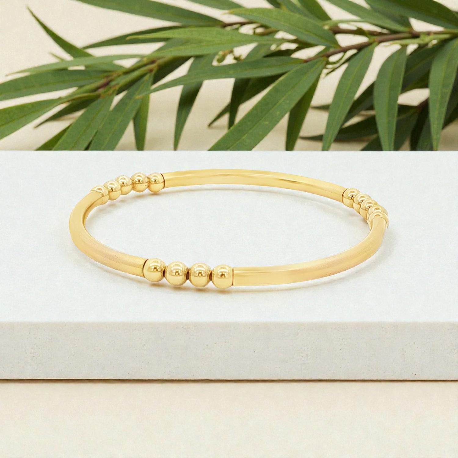 A gold-colored bracelet with a row of small, round beads on it, placed on a white surface with green leaves in the background.