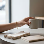 A hand holding a white mug is resting on a table, with a folded napkin and a notebook nearby.