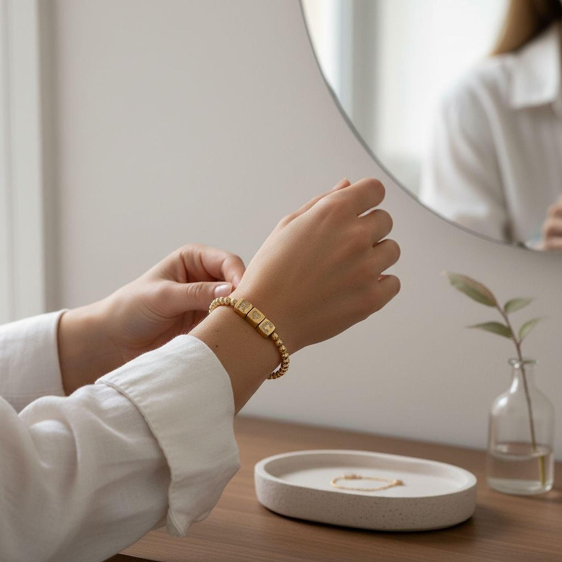 A person's hands adjusting a gold bracelet on a wooden surface, with a round mirror and a small vase with a plant in the background.