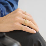 May include: A close-up of a hand wearing three gold rings. The ring on the index finger is a delicate chain with a heart charm. The ring on the middle finger is a beaded ring with small, round gold beads. The ring on the ring finger is a simple gold band.