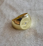 A gold ring with a large eye design on it, resting on a textured surface.
