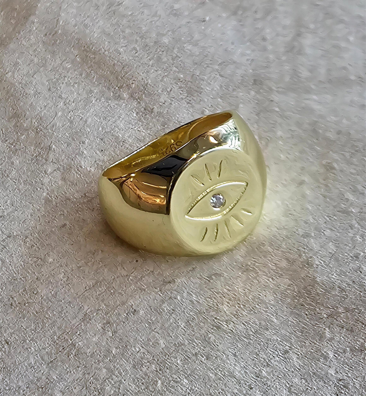 A gold ring with a large eye design on it, resting on a textured surface.